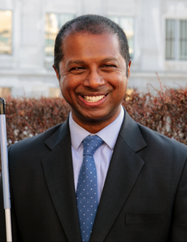 A blind man smiling, wearing a black suit with a blue tie and holding a cane.