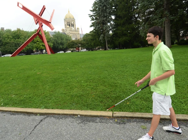 A young man walking with a cane, wearing a green shirt, white shorts and white shoes. He's walking in front of a park with a red sculpture.
