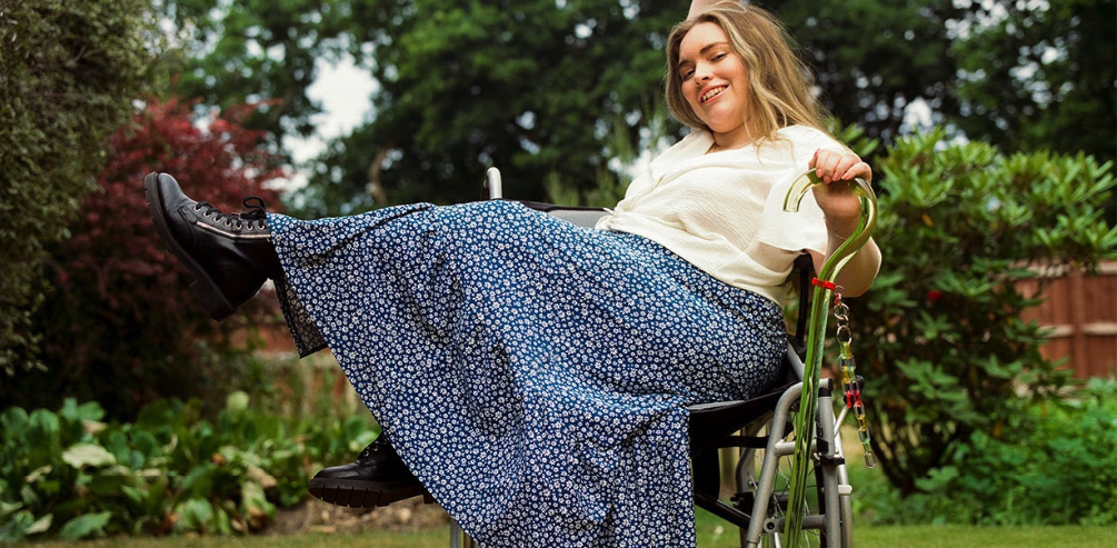 A blonde woman sitting on a wheelchair, wearing a white blouse, a floral, blue skirt and black boots. She's holding a transparent cane.